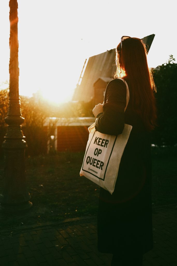 A person with long red hair and a long black coat, looking at an open bridge in the center of Amsterdam during sunset. They are carrying a "KEER OP QUEER" tote bag on their shoulder.